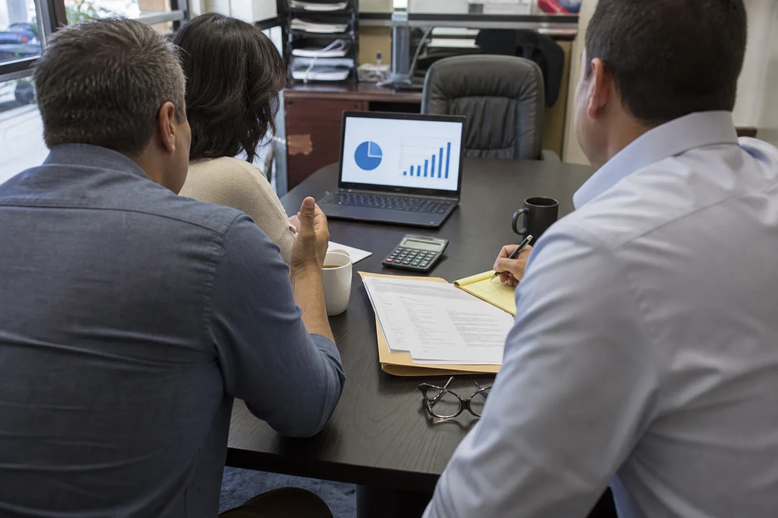 Over-the-shoulder view of an agent in a light blue shirt taking notes on a legal pad while a couple reviews charts on a laptop at a small office conference table.